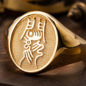 Puede incluir: Un anillo de sello dorado con una cara ovalada, un fondo texturizado y un carácter chino pulido en relieve en el centro. La banda es lisa y brillante, y el diseño general presenta un acabado en color oro metálico en toda la pieza.