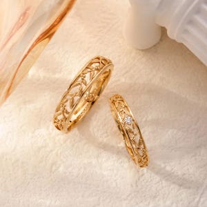 Puede incluir: Dos anillos de oro con un intrincado patrón de hojas y enredaderas en relieve a lo largo de las bandas. El anillo más pequeño incluye una única piedra preciosa redonda, pequeña y transparente, engastada en el centro del diseño.