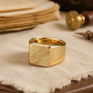 May include: A gold-toned signet ring featuring a flat, square face with an engraved dinosaur silhouette. The design includes textured, pebbled detailing on the surface surrounding the central figure, set against a polished, smooth band.