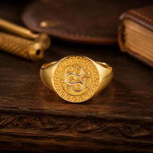 May include: A gold signet ring featuring a circular face with a textured dragon emblem at the centre, surrounded by a detailed Greek key border pattern. The ring has a polished finish and a smooth, rounded band that tapers towards the back.