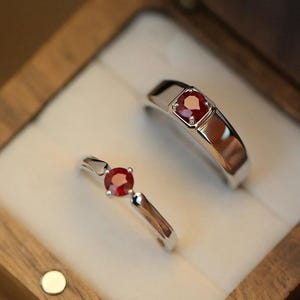 May include: Two silver-toned rings featuring round, faceted red gemstones. One ring has a wider band with a bezel-set stone, while the other features a thinner band with a prong-set stone. Both rings are displayed inside a wooden box.