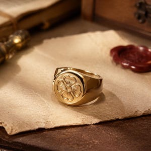 Può includere: Un anello a sigillo in oro lucido con una faccia circolare incisa con un design a fiore a quattro petali. L'anello presenta una fascia liscia e arrotondata e una finitura metallica riflettente, con un motivo floreale dettagliato al centro.