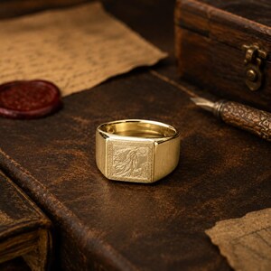 May include: A gold signet ring featuring a square face with an engraved illustration of a figure with flowing hair reaching toward a crescent moon, framed by a textured border. The band is smooth and polished, reflecting light on its surface.