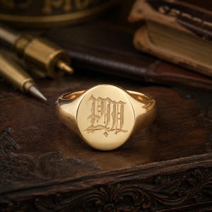 Puede incluir: Un anillo de sello de oro pulido con una cara circular que presenta una letra M gótica grabada. El anillo tiene una banda lisa y redondeada con un acabado metálico reflectante que destaca los detalles de la caligrafía.