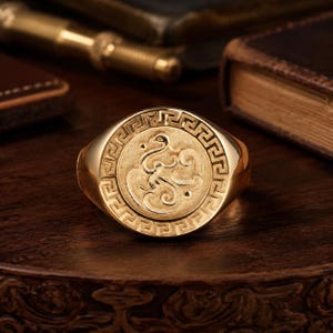 May include: A gold signet ring featuring a circular face with an engraved mythical creature at the center, surrounded by a detailed Greek key pattern border. The polished gold band has a smooth finish and a rounded profile, showcasing intricate metalwork.
