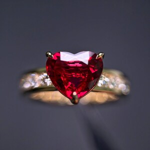 May include: A gold ring featuring a large, faceted, heart-shaped red gemstone as the centrepiece. The band of the ring is embellished with small, sparkling white stones that are slightly out of focus, highlighting the vibrant red heart stone.