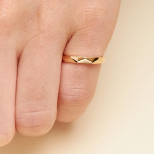 May include: A gold ring featuring a polished finish and a distinct geometric, faceted design on the front surface. The band has a smooth, uniform width and a reflective metallic surface that catches the light, creating sharp angles and highlights.
