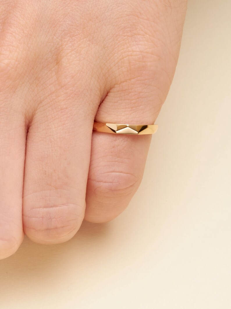 May include: A gold ring featuring a polished finish and a distinct geometric, faceted design on the front surface. The band has a smooth, uniform width and a reflective metallic surface that catches the light, creating sharp angles and highlights.