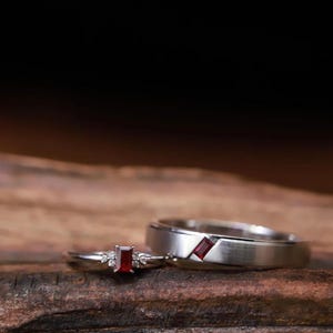 May include: A pair of silver-toned rings. One ring features a central rectangular red gemstone flanked by small clear stones. The second ring is a wider band with a brushed finish, inlaid with a single rectangular red gemstone set flush.