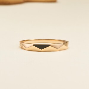 May include: A gold ring featuring a faceted geometric band design. The surface of the ring displays alternating smooth gold facets and a single black triangular inlay, with a small section of the band accented by tiny embedded clear stones.