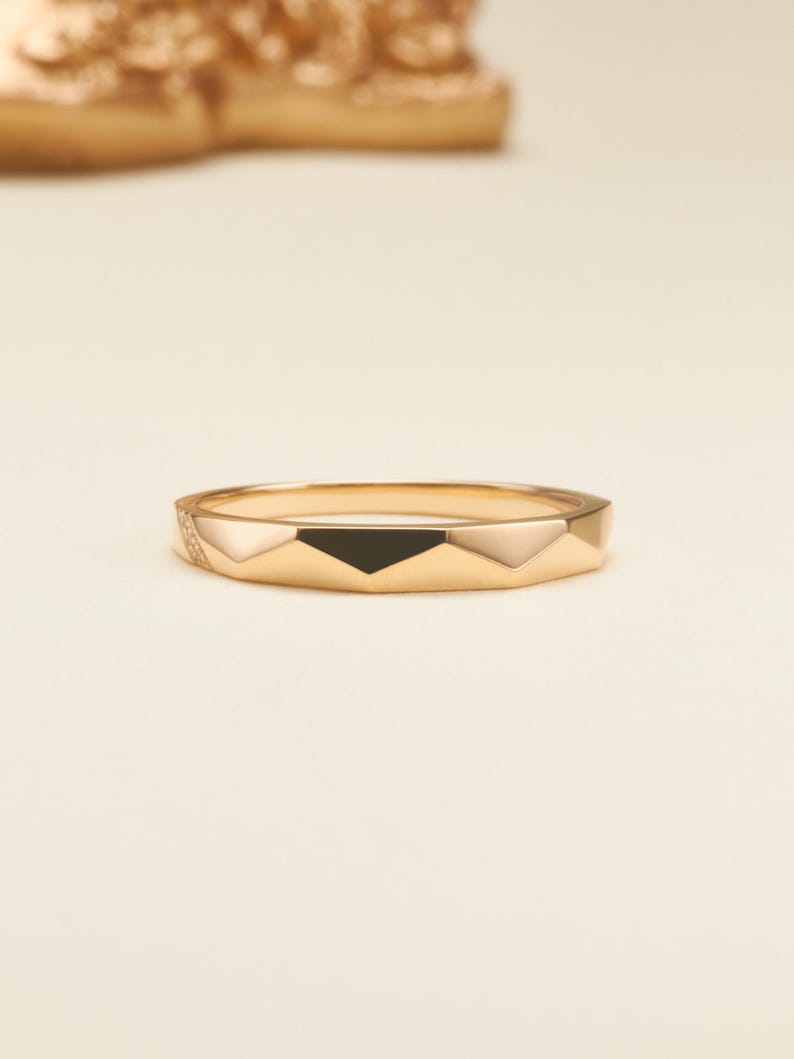 May include: A gold ring featuring a faceted geometric band design. The surface of the ring displays alternating smooth gold facets and a single black triangular inlay, with a small section of the band accented by tiny embedded clear stones.