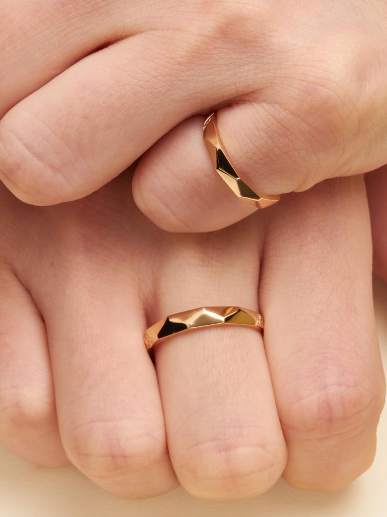 May include: Two polished gold rings with a faceted, geometric surface design. Each ring features a series of angular, flat planes that create a multi-faceted appearance, reflecting light across the metallic surface of the bands.