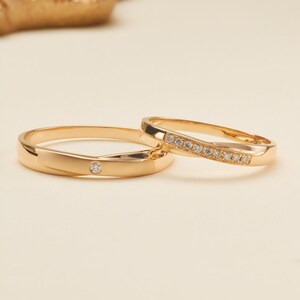 Könnte beinhalten: Zwei goldene Ringe mit polierter Oberfläche. Ein Ring ist mit einem einzelnen Diamanten im Rundschliff in der Mitte besetzt. Der zweite Ring hat ein geschwungenes Design, das mit einer Reihe von sieben kleinen Diamanten verziert ist.
