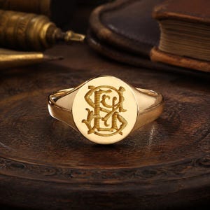 May include: A gold signet ring featuring a polished, smooth band and a flat, circular face. The center of the ring is engraved with an intricate, stylized monogram consisting of the intertwined letters S and G in a decorative, calligraphic font.