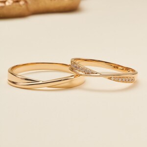 May include: Two gold rings with a polished finish and a twisted, crossover design. One ring features a smooth, plain band, while the other ring is accented with a row of small, clear, sparkling stones set along the twisted section of the band.