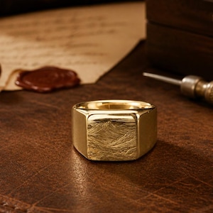 May include: A gold signet ring featuring a flat, square face engraved with a detailed mountain landscape. The band is smooth and polished, with a high-shine finish that reflects light across its surface, highlighting the intricate texture of the mountain scene.