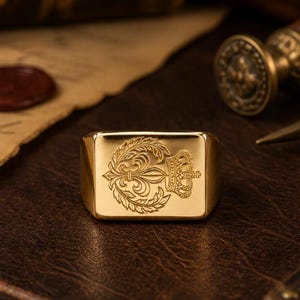 May include: A gold-toned rectangular signet ring featuring an engraved design on its flat face. The engraving depicts a central fleur-de-lis symbol flanked by leafy laurel branches, with a detailed crown positioned above the floral emblem.