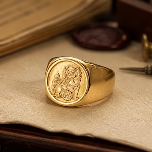 Pode incluir: Um anel de sinete dourado com uma face circular que apresenta um design em relevo de um leão guardião em pé com uma pata apoiada sobre uma esfera. O anel tem um acabamento polido e uma banda lisa e cónica que se une ao topo circular.