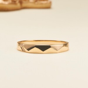 May include: A gold ring featuring a faceted geometric band design with sharp angular edges. One side of the band includes a small section embedded with tiny clear stones, while the rest of the surface displays a polished metallic finish.