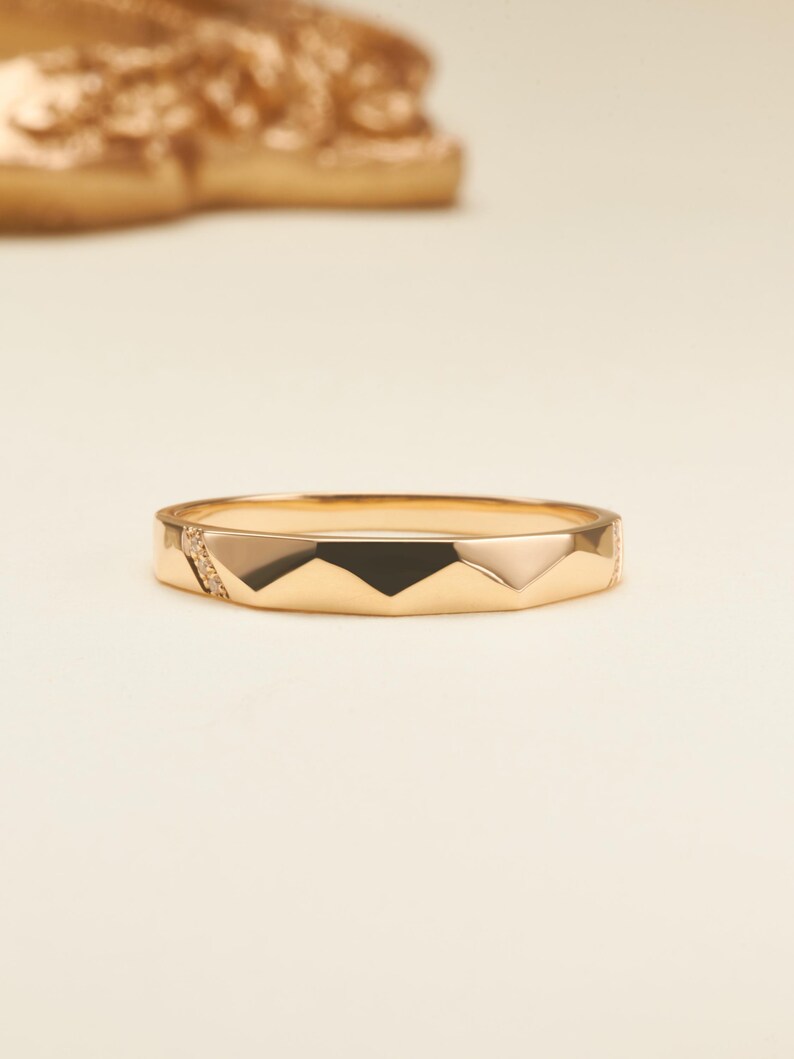 May include: A gold ring featuring a faceted geometric band design with sharp angular edges. One side of the band includes a small section embedded with tiny clear stones, while the rest of the surface displays a polished metallic finish.