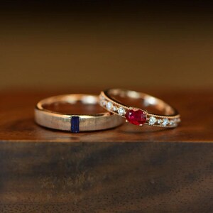 May include: A brushed silver-tone band featuring a single rectangular blue sapphire and a rose gold-tone band set with a central oval red ruby flanked by small round white diamonds along the band, showcasing a delicate beaded edge detail.
