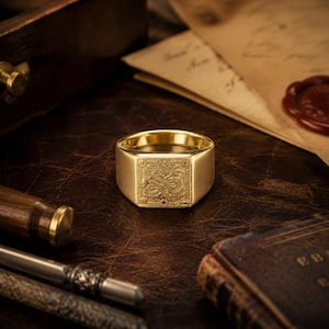 Puede incluir: Un anillo de sello de oro con una cara cuadrada que presenta un intrincado diseño de dragón grabado. El anillo tiene una banda lisa y pulida, mostrando un trabajo detallado en metal de un dragón enroscado dentro de un marco cuadrado.