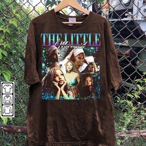 May include: Brown t-shirt featuring a graphic of characters from "The Little Mermaid" movie. The design includes the movie title in a gradient font and images of the characters. The shirt is hanging on a wooden hanger.
