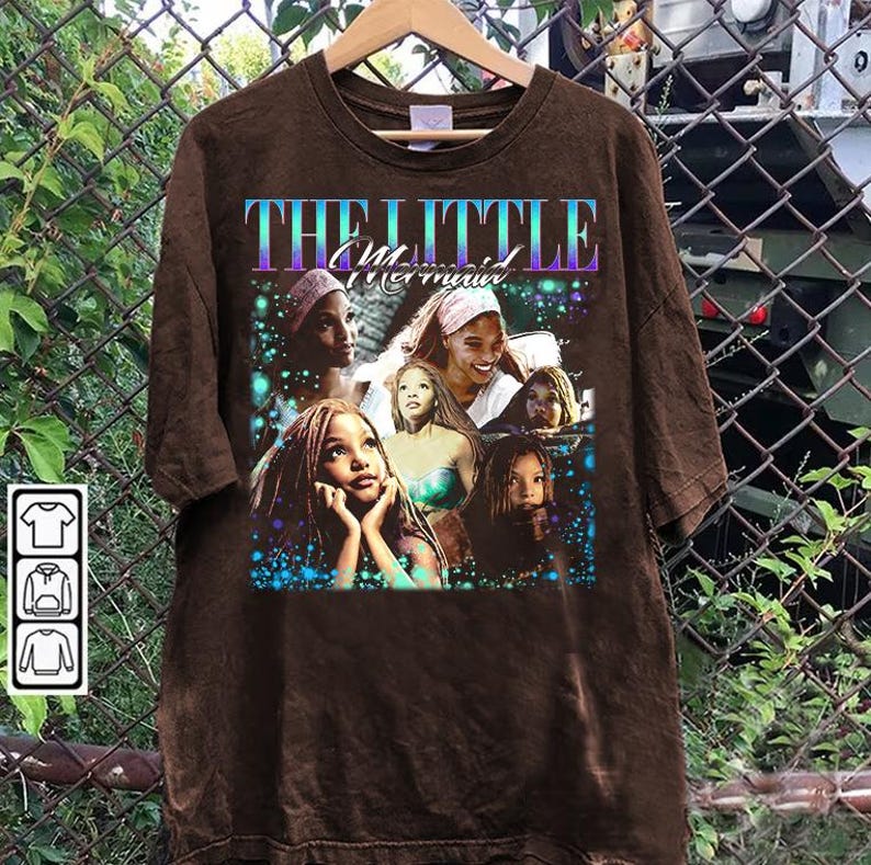 May include: Brown t-shirt featuring a graphic of characters from "The Little Mermaid" movie. The design includes the movie title in a gradient font and images of the characters. The shirt is hanging on a wooden hanger.