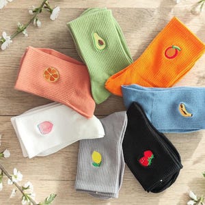 Könnte beinhalten: Eine Sammlung bunter Socken, jede mit einer anderen gestickten Frucht verziert. Die Socken sind in den Farbtönen Orange, Grün, Blau, Weiß, Grau und Schwarz gehalten und zeigen Designs wie Avocado, Orangenscheibe, Pfirsich, Zitrone, Banane und Kirschen.