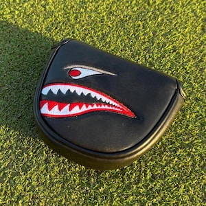 Könnte beinhalten: Schwarze Golfschlägerhaube mit einem Haifischmaul-Design. Die Haube hat einen schwarzen Hintergrund mit einer weißen, roten und schwarzen Haifischmaul-Grafik. Sie liegt auf einer grünen, grasbewachsenen Oberfläche.