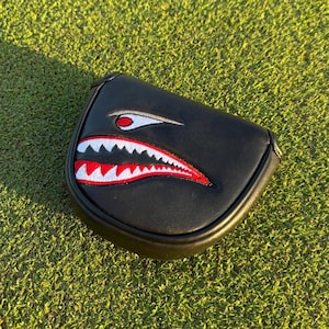 May include: Black golf club head cover with a shark mouth design. The cover features a black background with a white, red, and black shark mouth graphic. The cover is lying on a green grassy surface.