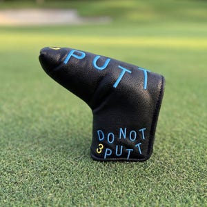Puede incluir: Funda negra para la cabeza del palo de golf con la palabra "PUTT" bordada en azul claro y "DO NOT 3 PUTT" en azul claro y amarillo. La funda está en un campo de golf verde.