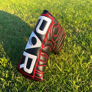 May include: A black golf putter headcover featuring red stitched contour lines and large white embroidered letters spelling out D-U-R. The design includes a small white circular emblem with a black golf ball graphic on the side.