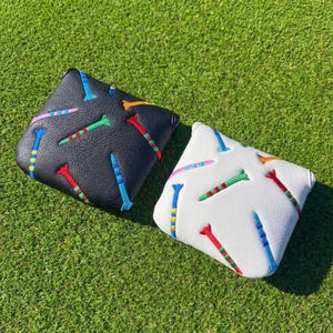 May include: Two golf club head covers. One is black, the other is white. Both have a pattern of colourful golf tees. The tees are red, green, blue, orange, and pink. The covers are on a green grass background.