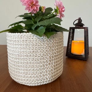 May include: A cream-colored, crocheted plant pot holding a flowering plant with pink blooms and green leaves. A decorative, dark metal lantern with a lit candle sits to the right. The pot is on a wooden surface.