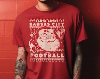 May include: Red t-shirt with a white graphic of Santa Claus holding a football and a beer. The graphic says "Santa Loves Kansas City Football" with a "KC" logo.
