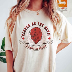 May include: A white t-shirt featuring a red devil graphic and the text "Clever as the Devil & Twice as Pretty".