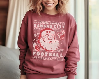 May include: A maroon sweatshirt with a white Christmas-themed design featuring Santa Claus wearing a red hat with the letters "KC" on it. The design also includes a football, snowflakes, and the text "Santa Loves Kansas City Football".
