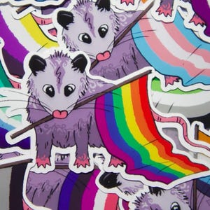 May include: A collection of stickers featuring cartoon opossums holding various pride flags. The opossums are purple with black ears and pink tongues. The flags include the rainbow pride flag, transgender flag, and others.