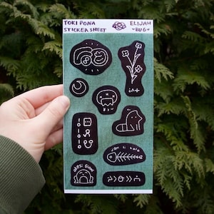 May include: A sticker sheet featuring various black and white illustrations, including a moon, a flower, and a fish skeleton. The text "TOKI PONA STICKER SHEET" is at the top. The sheet is held against a green background.