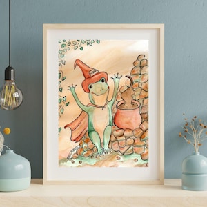 May include: A framed watercolor illustration of a green frog wearing an orange wizard hat and cape, standing next to a bubbling orange cauldron surrounded by brown stones. The hat features the text "HERE" written in black ink on the front.