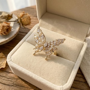 May include: A gold-coloured butterfly ring, set with clear and pearl-like stones, presented in a beige jewellery box. The butterfly design features detailed wings with various stone shapes and sizes, creating a sparkling effect. A delicate and elegant piece of jewellery.