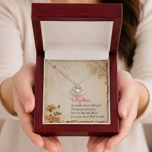 May include: A silver necklace with a heart-shaped pendant, presented in a dark red wooden box. The pendant has a clear stone in the center, surrounded by a rose gold and silver design. The box contains a card with the text "To My Mom".