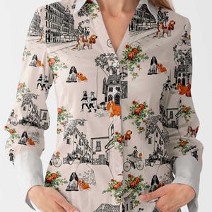 Puede incluir: Camisa abotonada color crema con un estampado caprichoso de perros, arreglos florales y bocetos arquitectónicos en blanco y negro. La camisa tiene cuello en V, mangas largas y puños y cuello blancos.