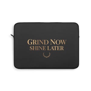 May include: Black laptop sleeve with the gold text "GRIND NOW SHINE LATER" and a gold laurel wreath. The sleeve has a zipper closure and is designed to protect a laptop.