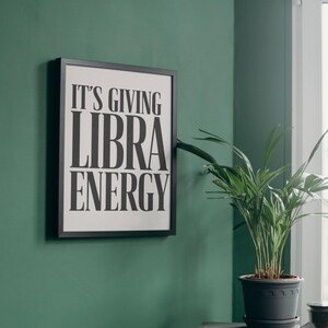May include: A framed print on a green wall reads "IT'S GIVING LIBRA ENERGY" in bold black letters. A potted plant sits on a small gray table next to the frame. The room has a modern, minimalist aesthetic.