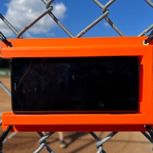 May include: A bright orange rectangular plastic smartphone mount secured to a chain-link fence with black elastic cords. The mount features a horizontal slot designed to hold a black smartphone securely in a landscape orientation for recording.