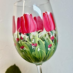 May include: A hand-painted wine glass with a design of pink tulips and green leaves. The tulips have white edges and are surrounded by small white dots.