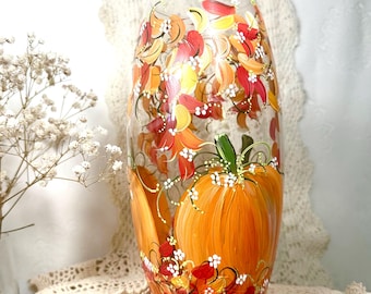 Puede incluir: Un jarrón de vidrio transparente con pintura a mano con temática otoñal. El jarrón presenta calabazas naranjas, hojas rojas, amarillas y naranjas, y pequeñas flores blancas. El diseño está pintado en el exterior del jarrón.