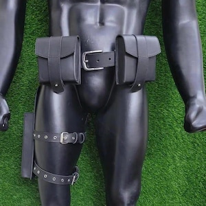 May include: Black leather utility belt with two pouches and a thigh strap. The belt features silver-toned buckles and rivets. The pouches have a rectangular shape with a flap closure. The thigh strap has multiple adjustment holes.